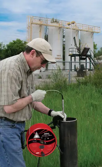 environmental technician wearing a beige cap and white gloves deploying a red solinst model 122 interface meter probe into a monitoring well casing with industrial storage tanks in the background